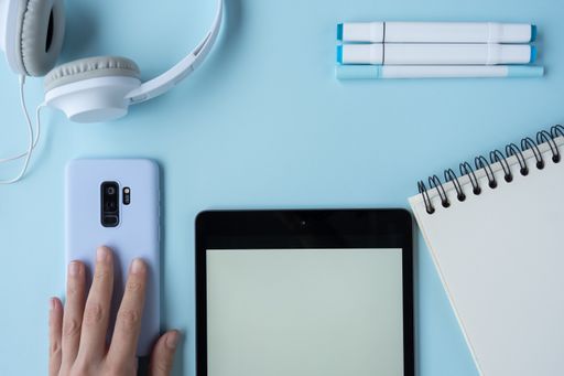 A high-angle shot of a person taking notes with a tablet, mobile phone, headphones, and markers on a blue desk