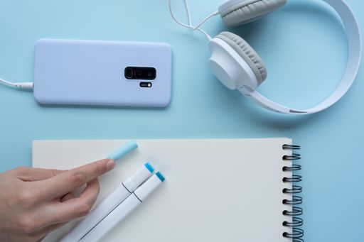 A high-angle shot of a person taking notes with a mobile phone, headphones, and markers on a blue desk