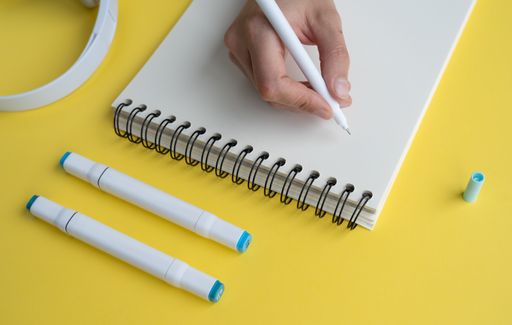 A person's hand writing in a notebook next to headphones on a yellow desk