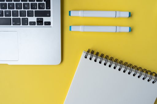 A high-angle shot of a yellow desk with a laptop, pens, and a notebook