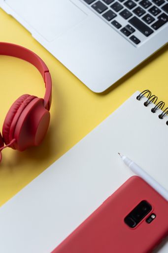 A vertical closeup shot of a notebook, a phone, pen headphones and a laptop on a yellow surface