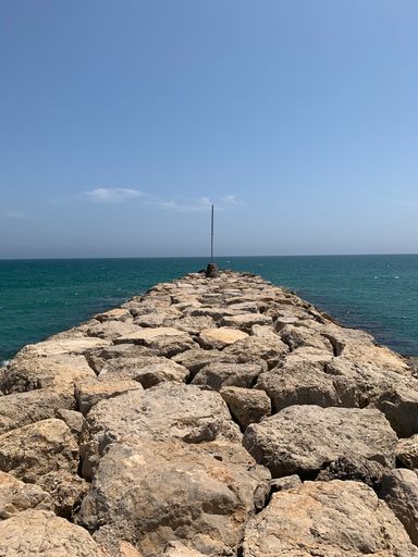 A vertical shot of a rocky jetty against the background of the teal sea and blue sky.