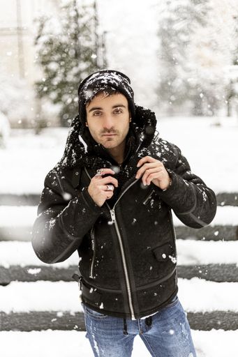 A vertical shot of a young man in black coat and hat standing in front of stairs in snowy winter