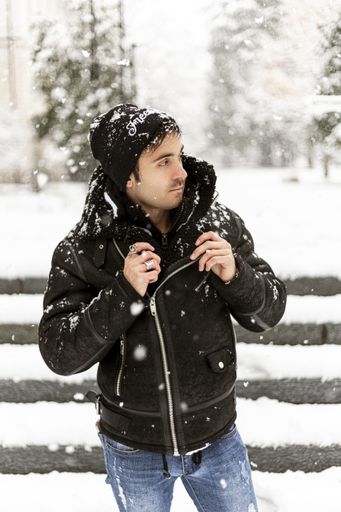 A vertical shot of a young man in black coat and hat standing in front of stairs in snowy winter