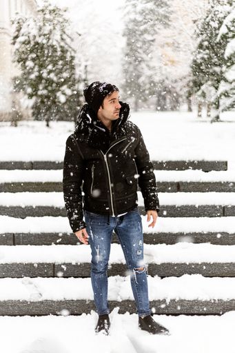 A vertical shot of a young man in black coat and hat standing in front of stairs in snowy winter