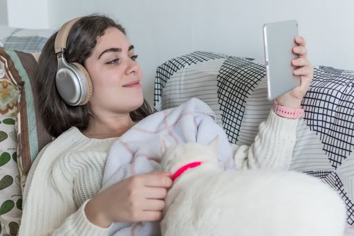 A closeup of a young female with headphones watching online content on a tablet lying on a sofa with her cat, in the living room at home
