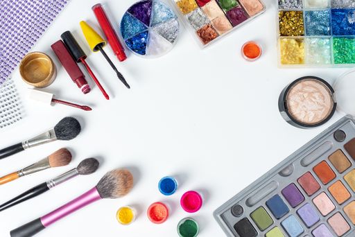 A high angle shot of brushes, highlighter, acrylic powder, eyeshadow, and watercolor palettes, mascara, lipsticks