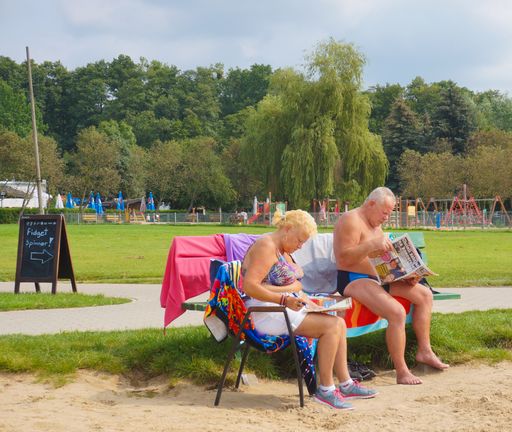 Man and woman in bathing clothes reading a magazine and newspaper while sitting on a wooden bench at a park.