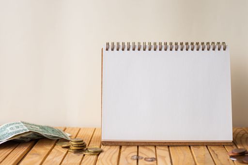 Empty Open Spiral Notebook Beside Stockpile Of Coins On Top Of Desk. Blank Opened Journal With Stacks Of Coin Placed On Wooden Table.