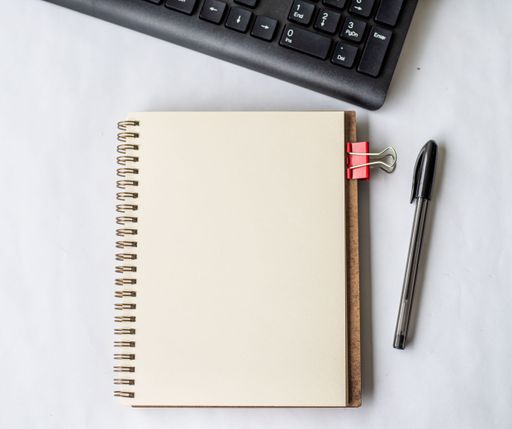 Empty Opened Clipped Journal Beside A Keyboard Pen On Top Of The Wooden Desk. Blank Open Notepad With Clip And Pencil Near Keypad Placed On Table.