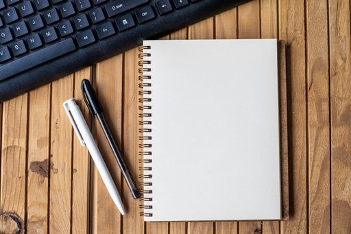 Empty Opened Clipped Journal Beside A Keyboard Pens On Top Of The Wooden Desk. Blank Open Notepad With Clip And Pencils Near Keypad Placed On Table.