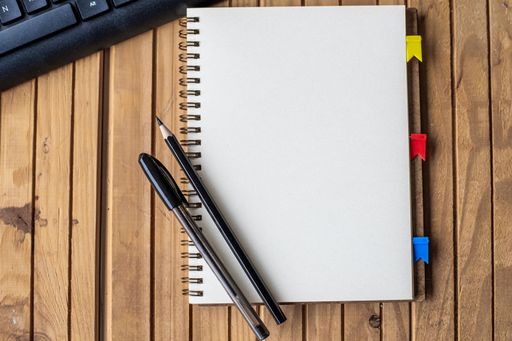 Empty Opened Clipped Journal Beside A Keyboard Pens On Top Of The Wooden Desk. Blank Open Notepad With Clip And Pencils Near Keypad Placed On Table.