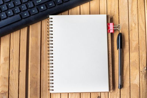 Empty Opened Clipped Journal Beside A Keyboard Pen On Top Of The Wooden Desk. Blank Open Notepad With Clip And Pencil Near Keypad Placed On Table.