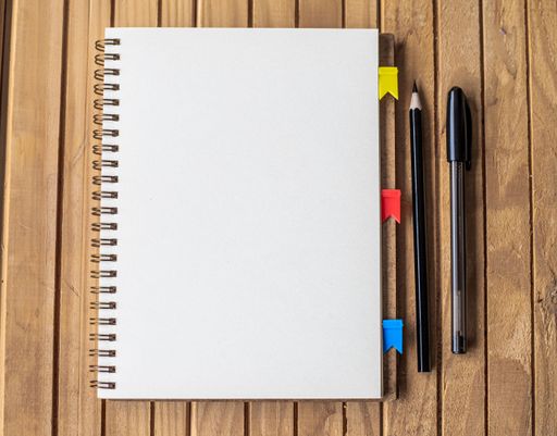 Empty Opened Clipped Journal Beside Pens On Top Of The Wooden Desk. Blank Open Notepad With Clip And Diffrent Pencils Placed On Table.