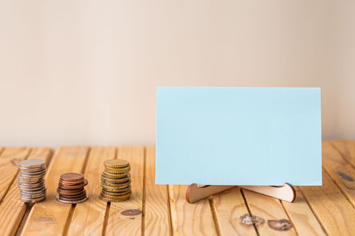 Empty Piece Of Paper On Holder Beside Stockpile Of Coins Over Desk. Blank Note Standing With Stacks Of Coin Placed On Wooden Table.