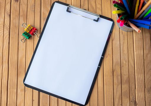 Plain Office Clipboard With Stack Of Pens In A Holder On Top Of A Table. Empty Paper Sheet With Colorful Pencils Container Placed On A Desk.