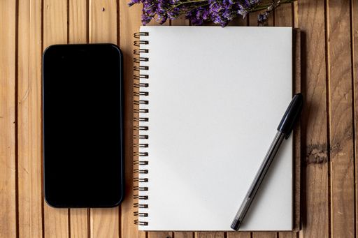 Empty Opened Journal Beside Mobile With Pens On Top Of Wooden Desk. Blank Spiral Notebook Pad Alongside A Phone With Ballpens Placed On A Table.