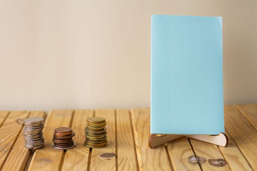 Empty Piece Of Paper On Holder Beside Stockpile Of Coins Over Desk. Blank Note Standing With Stacks Of Coin Placed On Wooden Table.