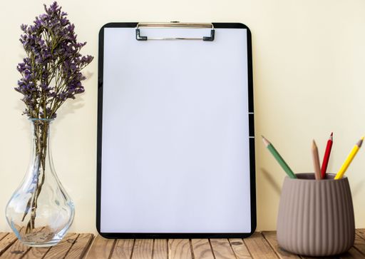Plain Office Clipboard With Stack Of Pens In A Holder Beside A Flower Vase. Empty Paper Sheet With Pencils Next To Floret Container Placed On Table.