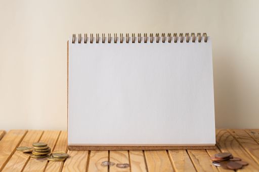 Empty Open Spiral Notebook Beside Stockpile Of Coins On Top Of Desk. Blank Opened Journal With Stacks Of Coin Placed On Wooden Table.