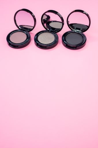 A vertical shot of black, pink, and gray single eyeshadow palettes on a pink surface