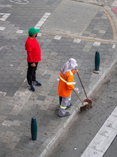 Medellin, Colombia - December 2 2020: Garbage Woman in Orange and Gray Uniform with a Hat is picking up Trash with a Broom and Dustpan on the Street