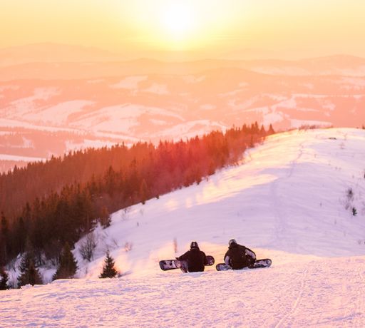 A high angle shot of two skiers sitting on the bright snow watching the sunset with a forest in the distance