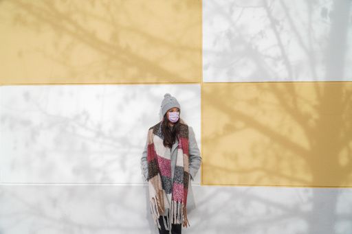 A young Caucasian female wearing a winter coat and a mask standing against a wall - the new normal due to Covid-19