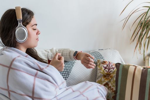 A young female with headphones drinking tea, resting on a sofa and looking at her watch