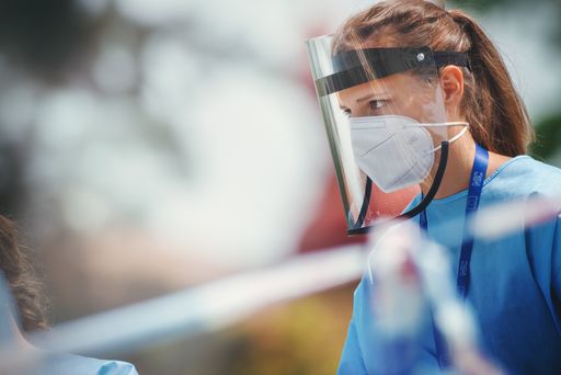 Female health care worker with a respirator and a plastic face shield standing on the street.