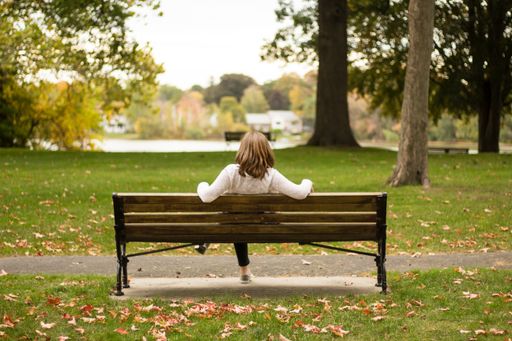 A female sitting on a bench in a park shot from behind in the fall with a lake and trees in the background