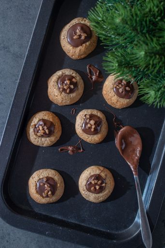 An assortment of freshly baked chocolate linzer cookies, sprinkled with nuts and topped with chocolate frosting, arranged on a black cookie tray