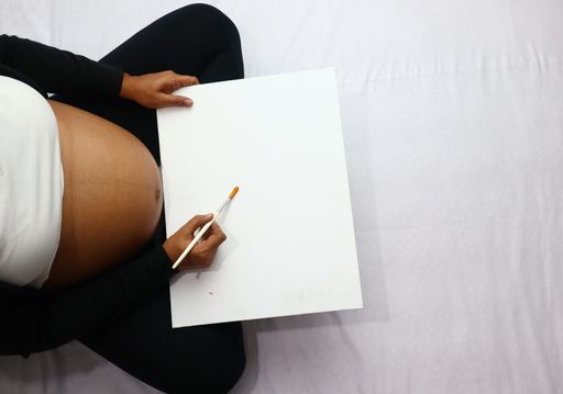 A top view of a young pregnant female painting on the bed at home