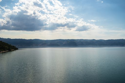 A sea surrounded by greenery and buildings with mountains under a cloudy sky on the background
