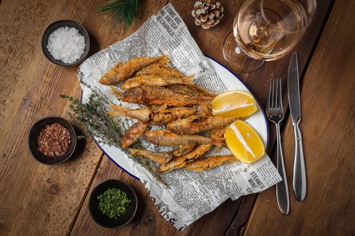A top view of small cooked fish with crusty topping, lemons and spices on a newspaper with wine