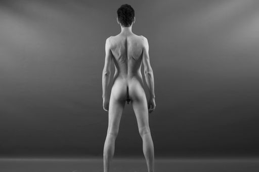 A grayscale shot of a slim male posing nude and showing his backside on a monochrome background