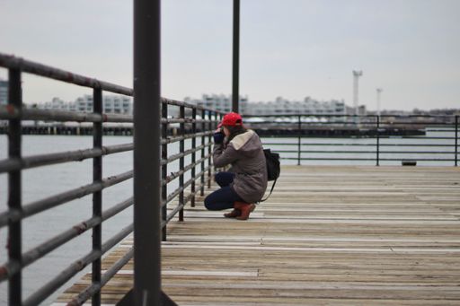 A female squatting down on a large wooden pier taking a photo of the ocean wearing winter cloths and a red baseball hat