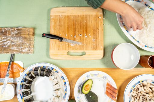 A top view shot of a cook cutting sushi on a cutting board with various ingredients on a table