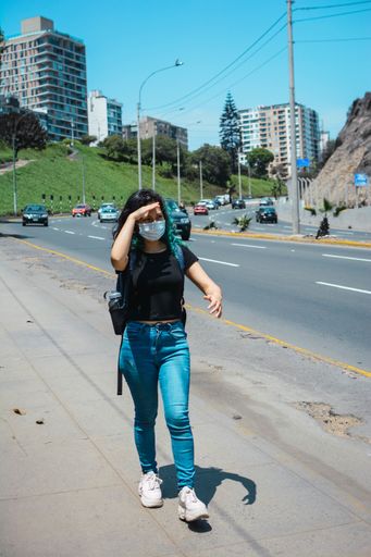 A vertical shot of a young Hispanic woman with a protective mask walking along the highway on a hot day