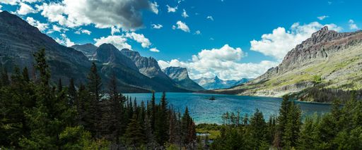 A wide shot of a lake surrounded by mountains and trees under a blue sky
