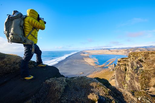 A male wearing a yellow jacket standing on a rock while taking a picture of the beautiful scenery