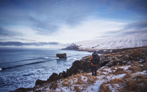 A male wearing a backpack and a jacket standing on the snowy hill while taking a picture of the beautiful sea