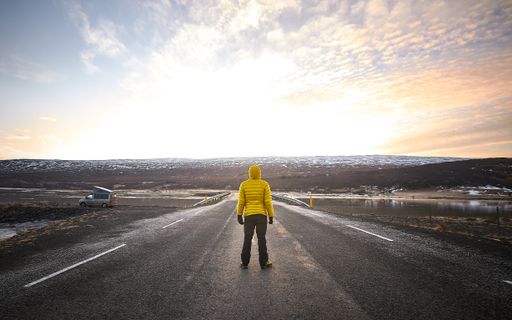 A male wearing a yellow jacket while standing in the middle of an empty road looking in the distance