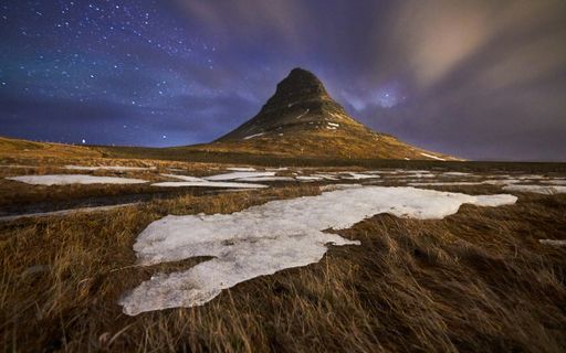 A beautiful shot of a dry grassy field with snow patches and a mountain in the distance under a star-filled sky
