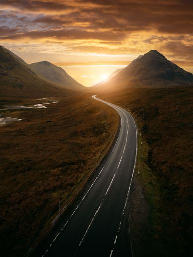 A scenic road winds through the mountains with a stunning sunset in the backdrop, providing a breathtaking view of the landscape