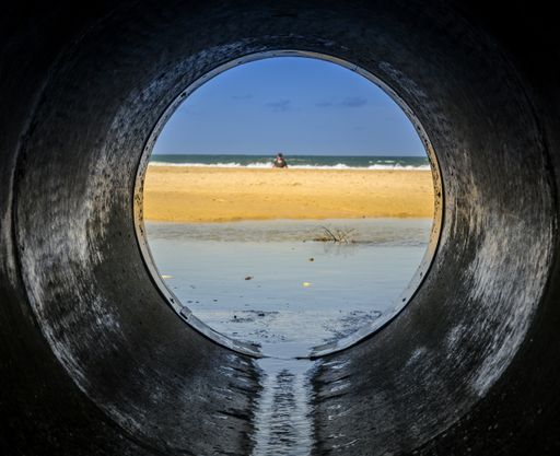 A look form a pipe looking at the beach surrounded by the sea with people in it under sunlight