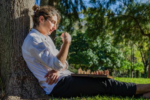 A shallow focus closeup of a male sitting under the tree while playing chess with a chessboard on his lap in the park