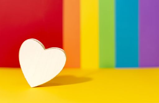 A selective focus shot of a white wooden heart on a background with the colors of the LGBT flag