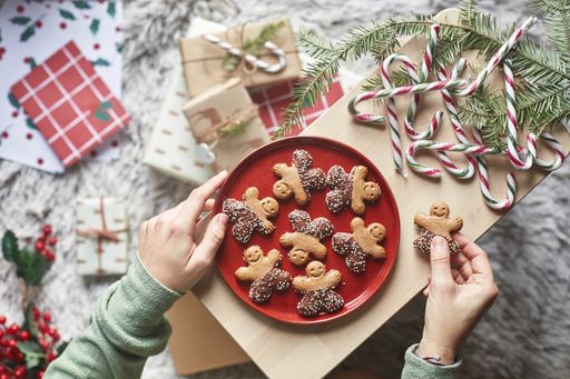 A top view of hands putting a gingerbread cookie on a red plate on a wooden board with candy sticks