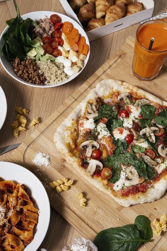 A vertical shot of an artisan pizza on the wooden board with smoothies, vegetables and pastries.